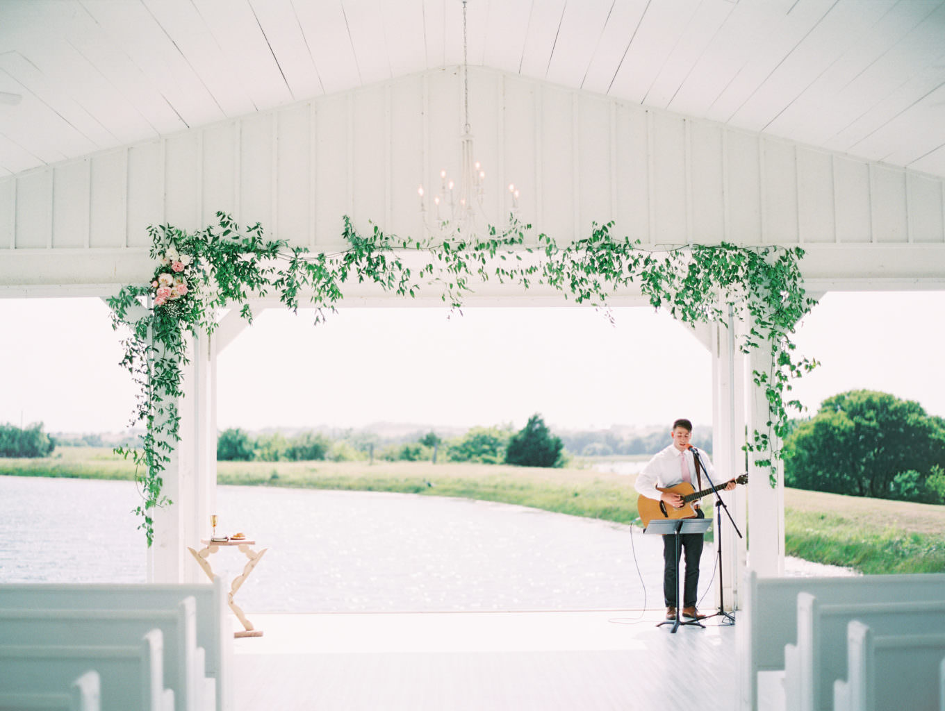 Musician with guitar at wedding ceremony set up, white chairs and green floral decor