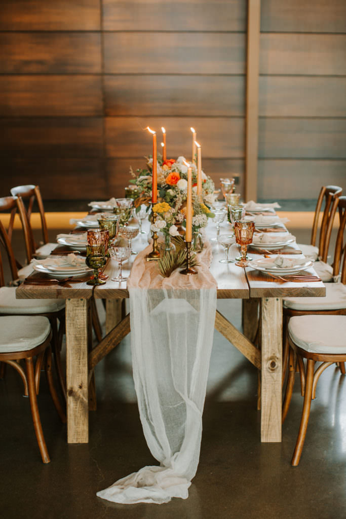 Boho styled wooden table with orange and peach colored candles, mixed florals, flatware, glasses and a tulle runner