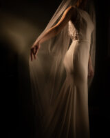 Moody dark image with a bride in the light holding wedding veil by Perla Images - New England photographers